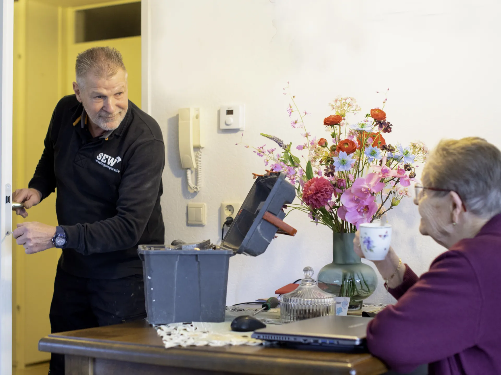 Monteur van Stichting Eelder Woningbouw repareert een deur terwijl een huurder toekijkt.