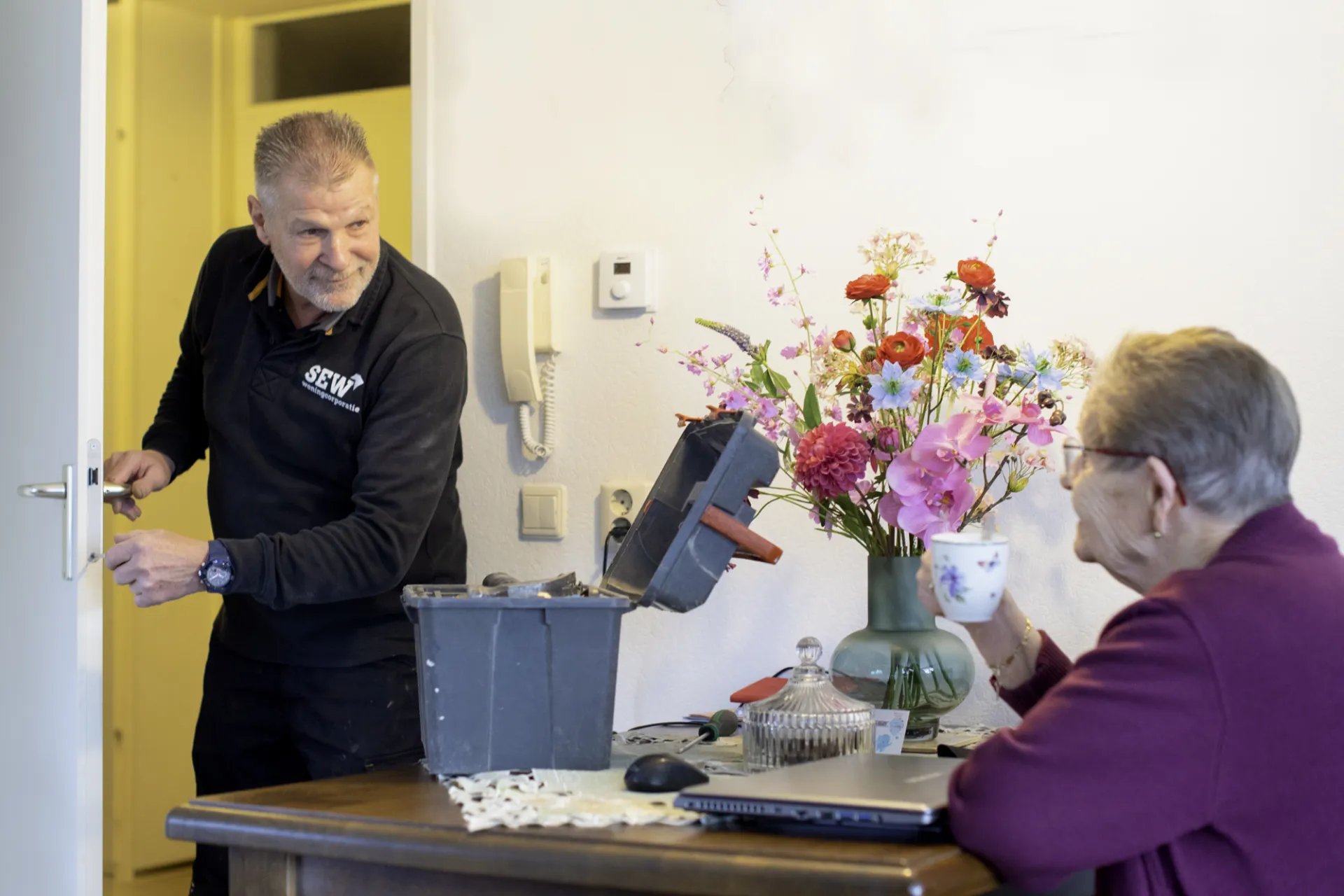 Monteur van Stichting Eelder Woningbouw repareert een deur terwijl een huurder toekijkt.